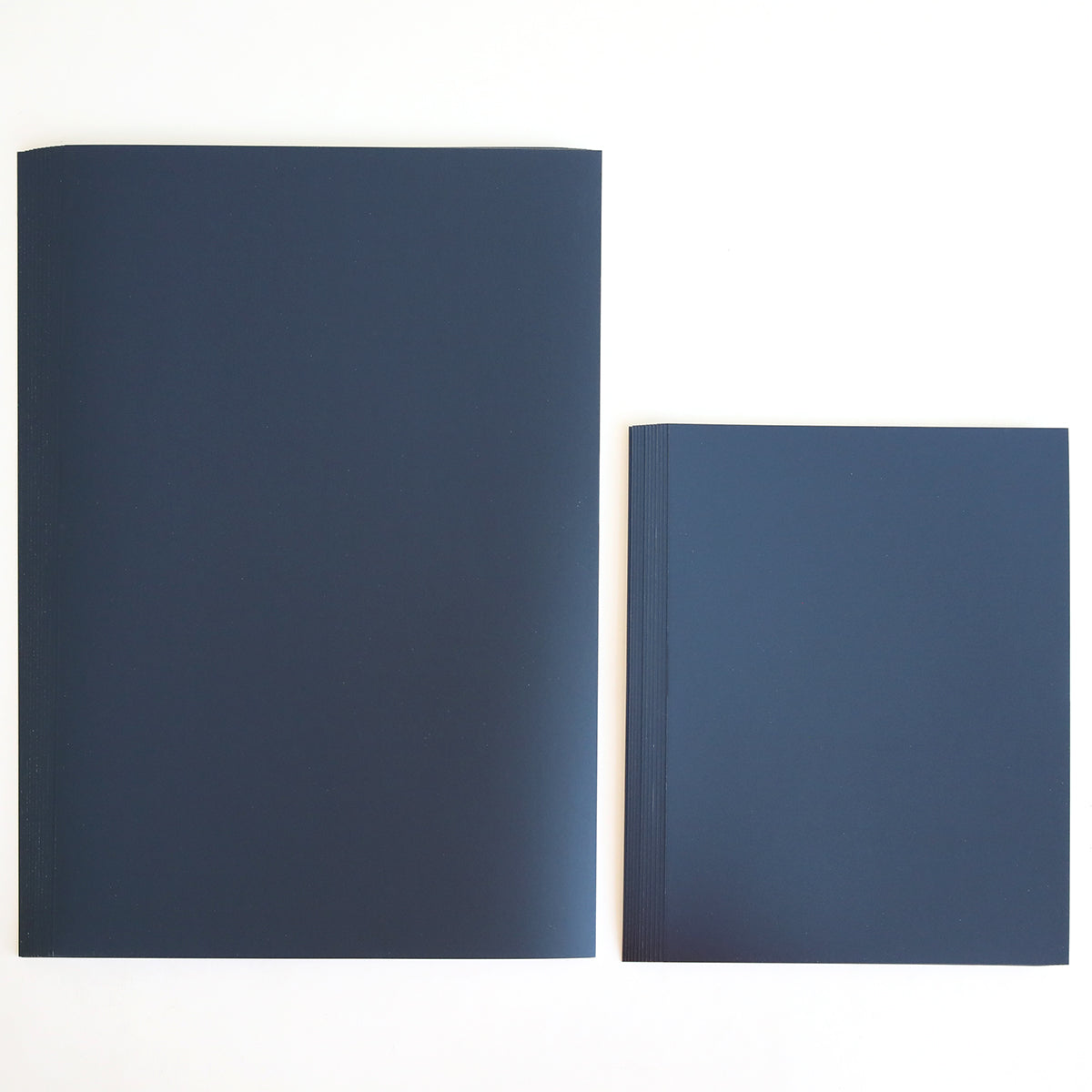 Two closed notebooks from the Celebration Foil Cardstock Collection, one larger and one smaller, in a dark blue shade are placed side by side on a plain white surface. Their sturdy covers resemble navy matte foil cardstock, making them perfect for stylish journaling or as inspiration for elegant paper crafts.