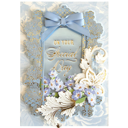 A decorative card featuring “On Your Special Day” in gold script, adorned with blue and white flowers, ornate borders, and a blue ribbon bow at the top against a floral-patterned background—perfect for conveying heartfelt birthday wishes. The elegant design is beautifully enhanced using the Set of 30 Gilded Small Sentiment Plates for an added touch of charm.
