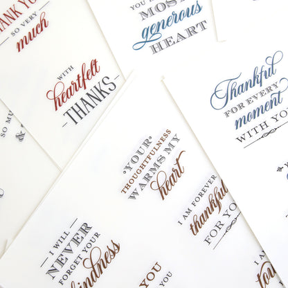A collection of white cards adorned with elegant, cursive thank you messages in black, blue, and red ink—each featuring heartfelt sentiments from the With Gratitude Sentiment Rub Ons—arranged in an overlapping display on a flat surface.
