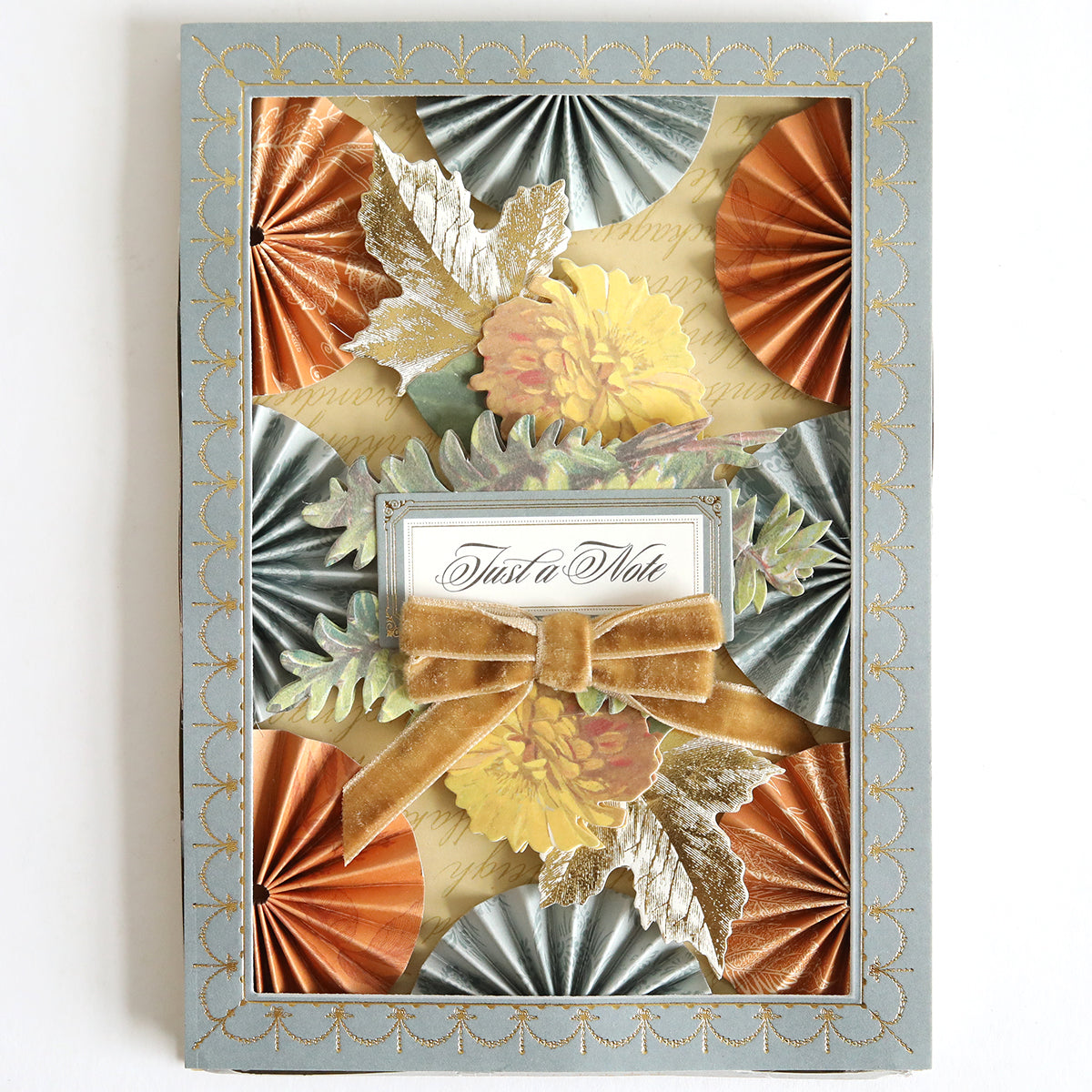 Introducing the Holiday Photo Mat and Card Layer Bundle: a stylish greeting card base featuring pleated metallic paper, elegant floral accents, a gold velvet bow, and a "Just a Note" cursive label, all beautifully framed with a patterned light blue border—ideal for card making or scrapbooking projects.