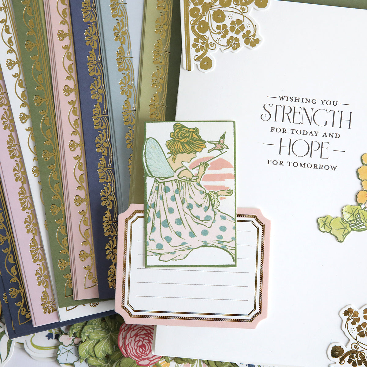 Featuring enchanting fairy artwork, the Dahlia Ultimate Papercrafting Collection includes notecards adorned with fairies, layered over beautifully patterned papers and a white card inscribed in gold and black with the message “Wishing you strength for today and hope for tomorrow,” making this collection ideal for creating handmade cards or bringing a magical touch to scrapbook projects.