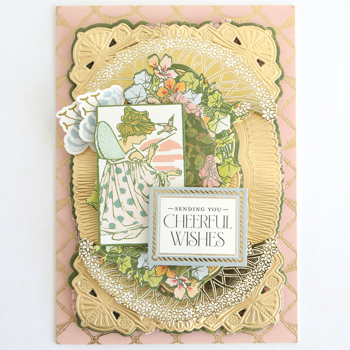 Add a touch of magic to your handmade cards or scrapbook pages with the Dahlia Ultimate Papercrafting Collection, featuring greeting cards adorned with gold decorative borders, an enchanting illustration of a fairy surrounded by flowers and birds, and a central message that reads “Sending You Cheerful Wishes” on a beautiful pink and gold patterned background.