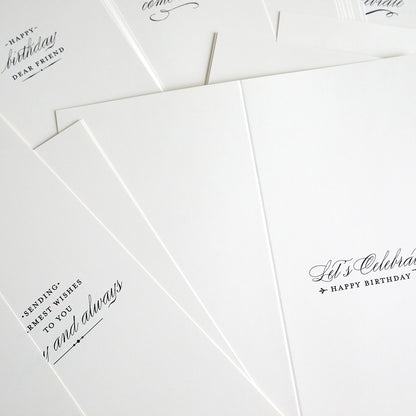 A close-up of Slimline Birthday Card Sentiment Inserts: several white cards with black cursive text and elegant ivory inserts, partially overlapping on a flat surface.