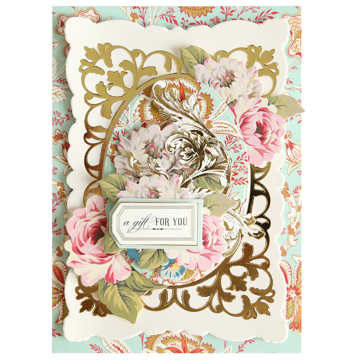 An ornate greeting card features pink and white flowers, gold embossing, and a label reading “a gift FOR YOU” on a decorative pastel floral background. The intricate design showcases a handmade touch, highlighted by an elegant frame created with the Oval Flourish Frame Gilded Hot Foil Plate.