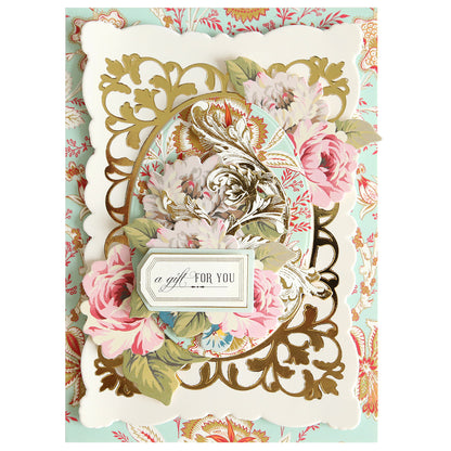 An ornate greeting card features pink and white flowers, gold embossing, and a label reading “a gift FOR YOU” on a decorative pastel floral background. The intricate design showcases a handmade touch, highlighted by an elegant frame created with the Oval Flourish Frame Gilded Hot Foil Plate.