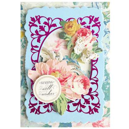 Decorative handmade card featuring the Oval Flourish Frame Gilded Hot Foil Plate with intricate floral cut-out details, layered pink and yellow flowers, a butterfly, and a round label reading “Sending Well Wishes.” The pastel floral patterned background adds an elegant touch, making it perfect for your handmade cards.