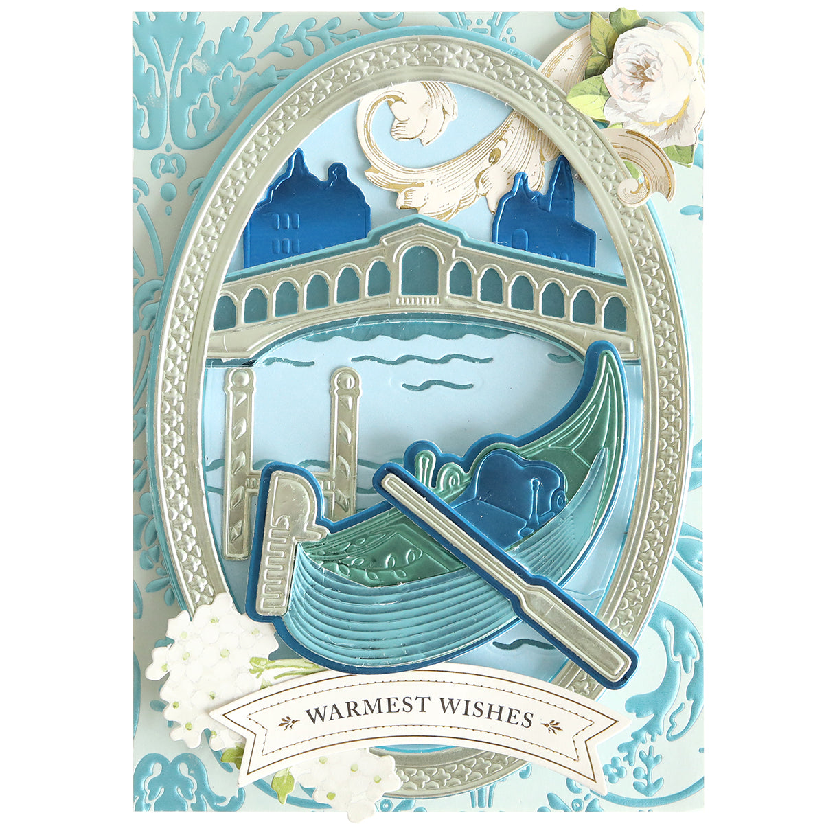 Greeting card with a layered illustration of a gondola on a canal, blue buildings, ornate border, and floral accents. Showcasing dimensional die cuts created using the Gondola 3D Scene Dies, inspired by Venice paper crafting. Text reads “Warmest Wishes.”.