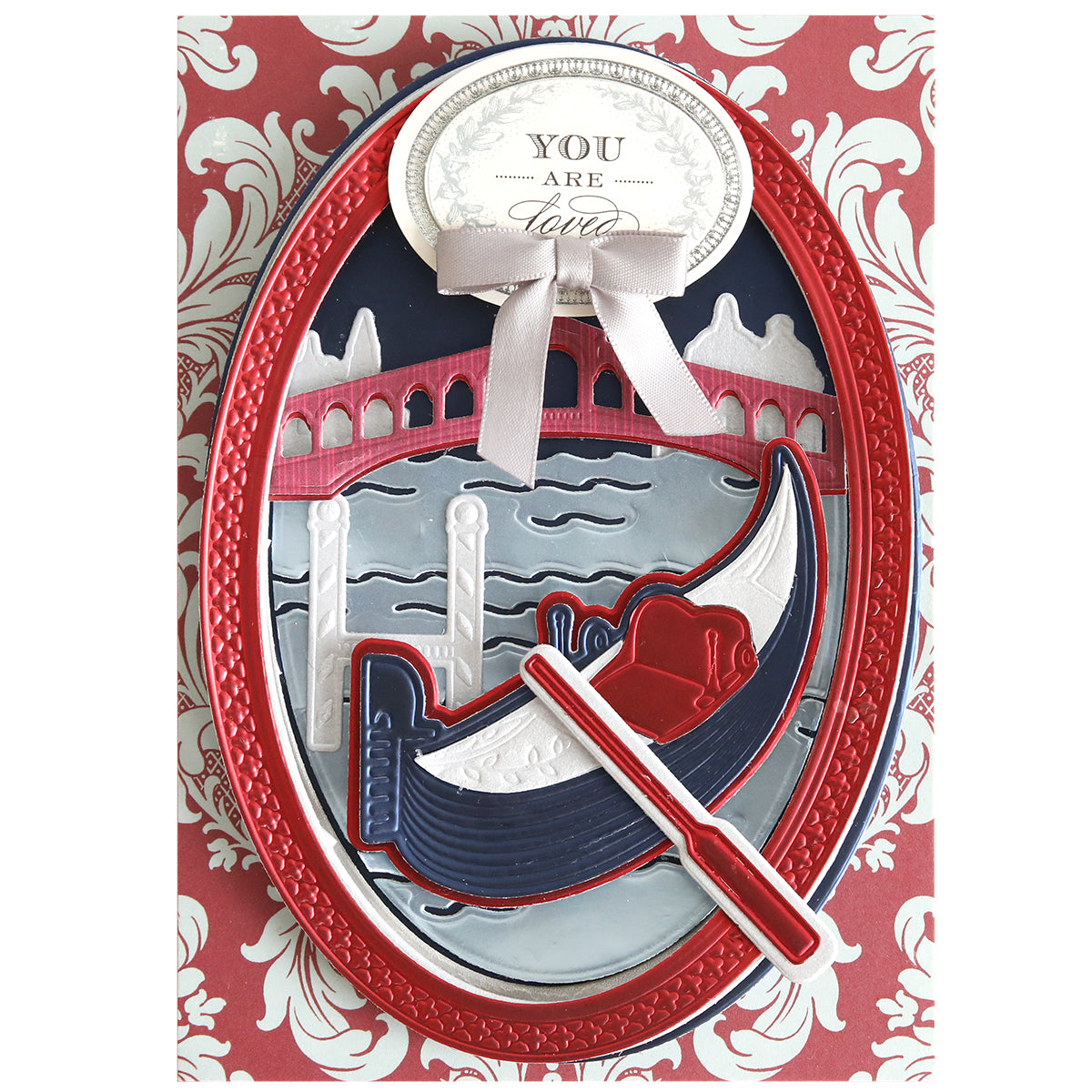 Greeting card with a red and white ornate border, featuring a gondola on a canal with a bridge, and a sign that says “You Are Loved” tied with a ribbon at the top—perfect for Venice paper crafting or for pairing with your Gondola 3D Scene Dies to create stunning dimensional die cuts.