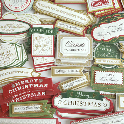 A variety of festive Christmas greeting cards and labels in red, green, and gold feature messages like “Merry Christmas” and “Season’s Greetings,” arranged in a scattered pattern. Some designs are decorated with charming 3D accents from the Favorite Christmas Sentiment Stickers collection, adding extra dimension and holiday cheer.