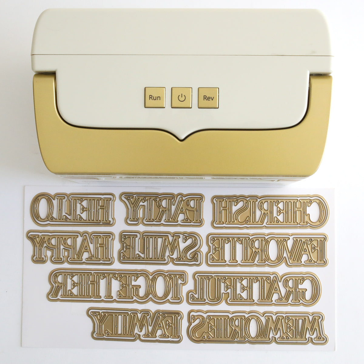 The Empress Mini Machine with Title Dies, featuring a gold and white design, is shown above the included reversed metal title dies laid out on a flat surface.