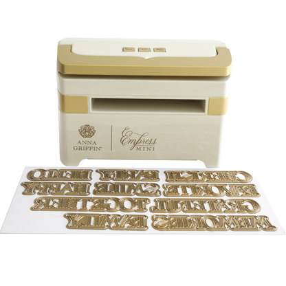 The Empress Mini Machine with Title Dies is shown, featuring a compact design and several gold metal title dies displayed in front.