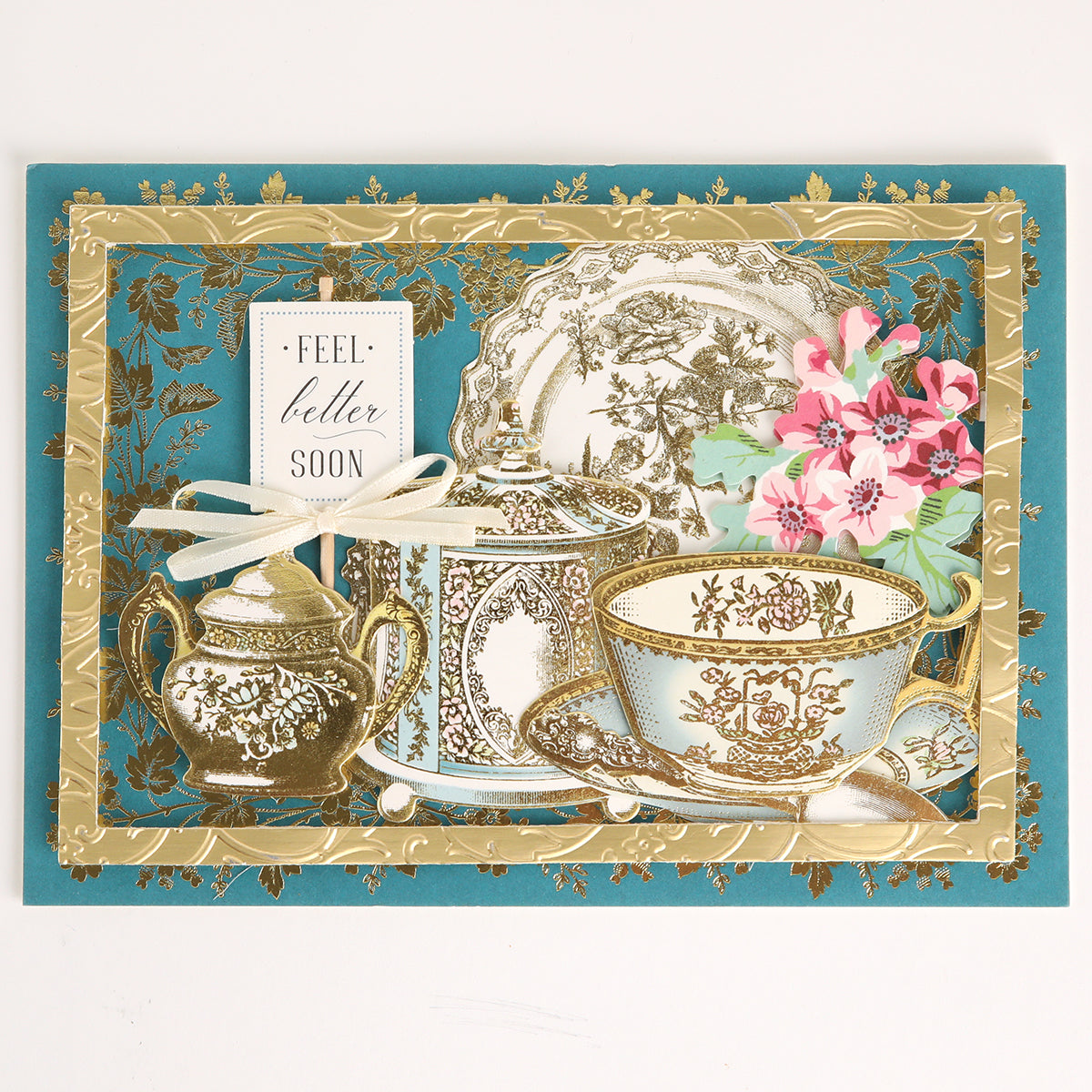 Handmade greeting card featuring ornate teacups, a teapot, and flowers on a blue and gold background. Decorated with accents from the Favorite Tea Embellishments 120 Count set, this card is finished with a white ribbon and a “Feel better soon” tag for an extra special touch.