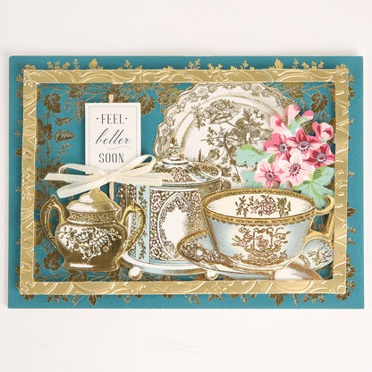Handmade greeting card featuring ornate teacups, a teapot, and flowers on a blue and gold background. Decorated with accents from the Favorite Tea Embellishments 120 Count set, this card is finished with a white ribbon and a “Feel better soon” tag for an extra special touch.