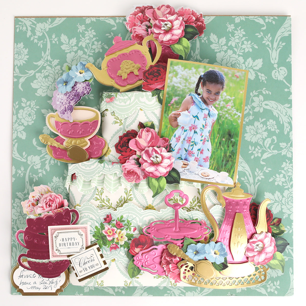 A scrapbook page with floral decor, teapots, teacups, and a photo of a young girl at a tea party, all arranged on a green patterned background and beautifully accented with Tea Party Cut and Emboss Dies for charming craft embellishments.