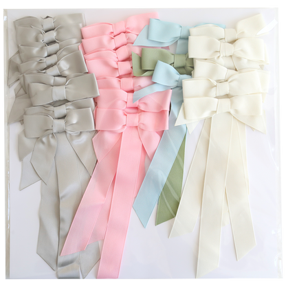 Seven large Pre-Tied Bows with Tails in light gray, pink, platinum, light green, light blue, and cream are arranged on a white surface.