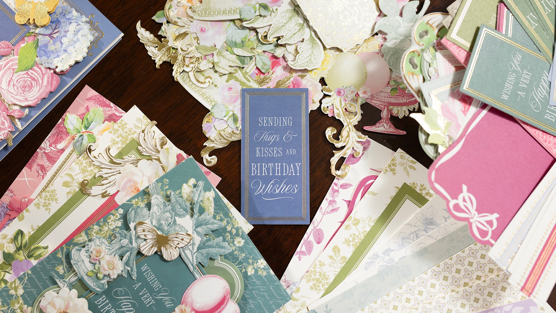 A variety of ornate, floral-themed birthday cards with decorative text and embellishments are arranged on a wooden surface.