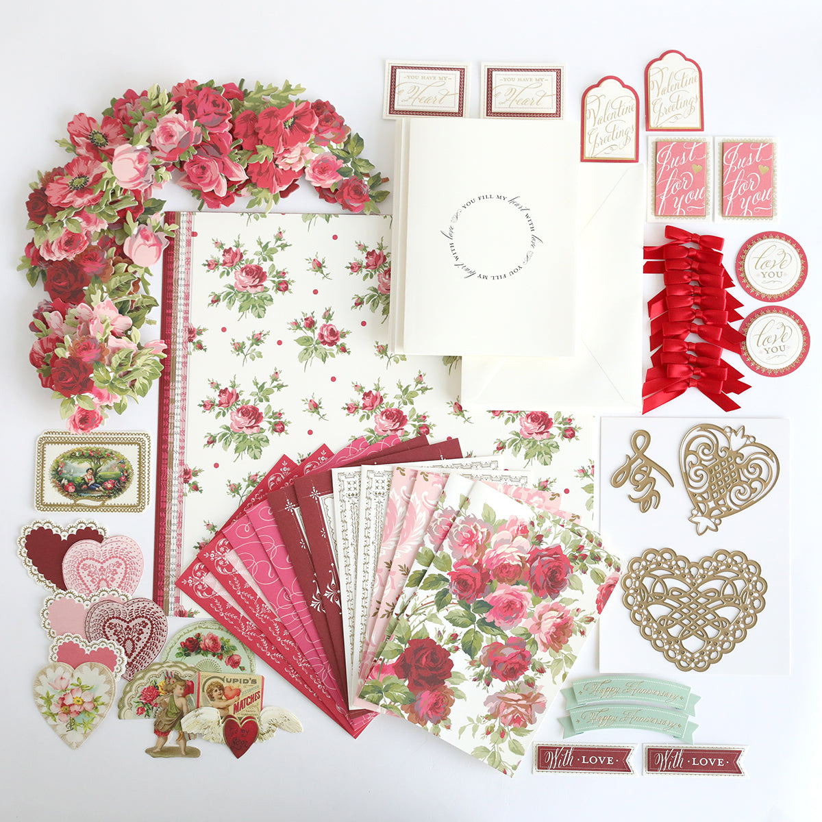 A flat lay of romantic-themed stationery, including floral papers and envelopes from the Anna Griffin® 2026 Papercrafting Mystery Box Subscription, plus heart-shaped cutouts, stickers, ribbons, and cards with love and rose designs.