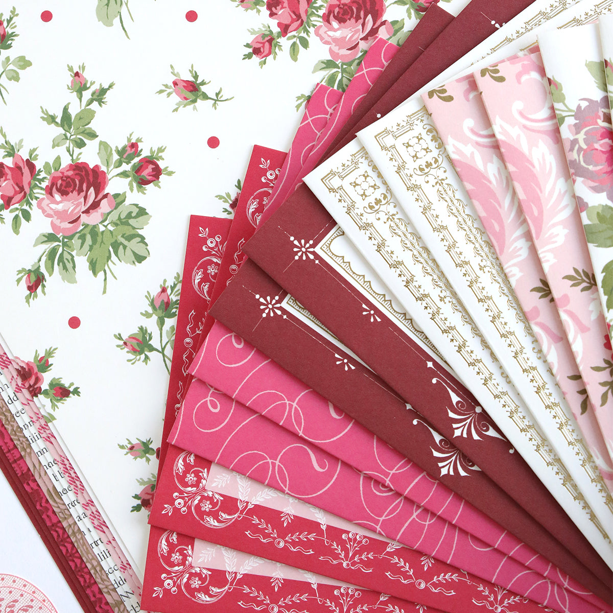 The Anna Griffin® 2026 Papercrafting Mystery Box Subscription includes decorative patterned papers in pink, red, and white, perfect for card making, arranged on a floral and polka dot background.