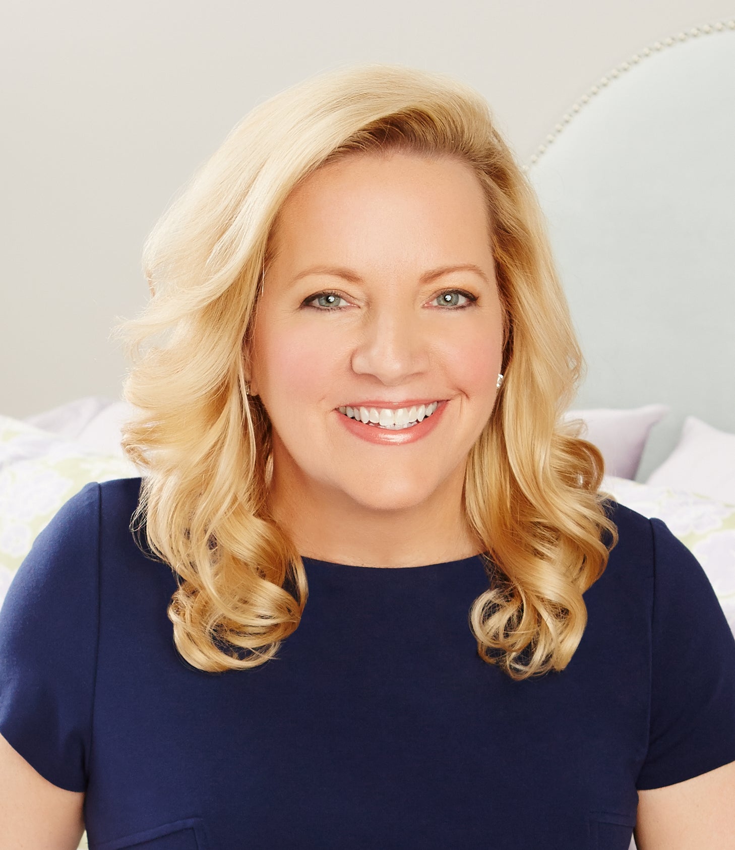 Smiling person with long blonde hair wearing a dark blue outfit against a light background.