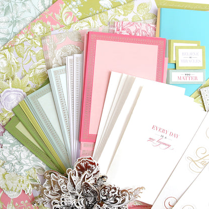 A display of decorative paper and envelopes in pastel colors, with floral patterns and motivational quotes. An ornate silver embellishment lies in the foreground.