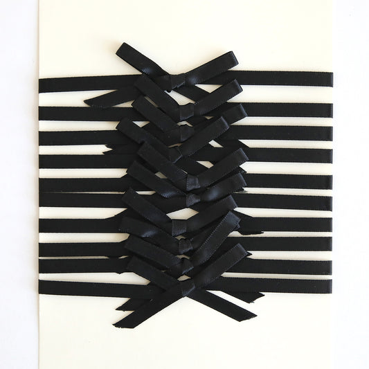 The Black Satin Bows 10 count features black ribbon embellishments arranged in a crisscross pattern on a white surface, forming elegant satin bows at the center.