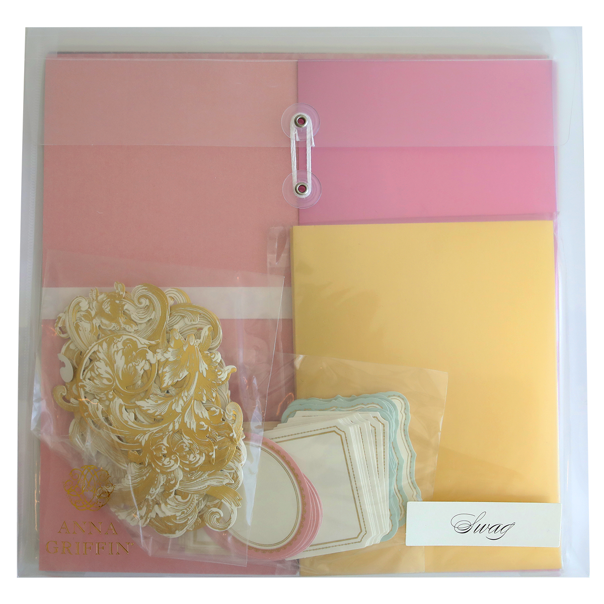 A clear plastic folder, perfect for scrapbooking or cardmaking, containing pink and yellow sheets, gold ornamental embellishments, white labels with pink and blue borders, and a small envelope labeled "Crop at Home II Swag." Ideal for crafting enthusiasts!