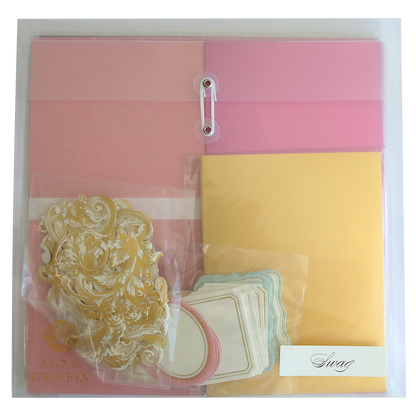 A clear plastic folder, perfect for scrapbooking or cardmaking, containing pink and yellow sheets, gold ornamental embellishments, white labels with pink and blue borders, and a small envelope labeled "Crop at Home II Swag." Ideal for crafting enthusiasts!