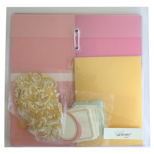 A clear plastic folder, perfect for scrapbooking or cardmaking, containing pink and yellow sheets, gold ornamental embellishments, white labels with pink and blue borders, and a small envelope labeled "Crop at Home II Swag." Ideal for crafting enthusiasts!