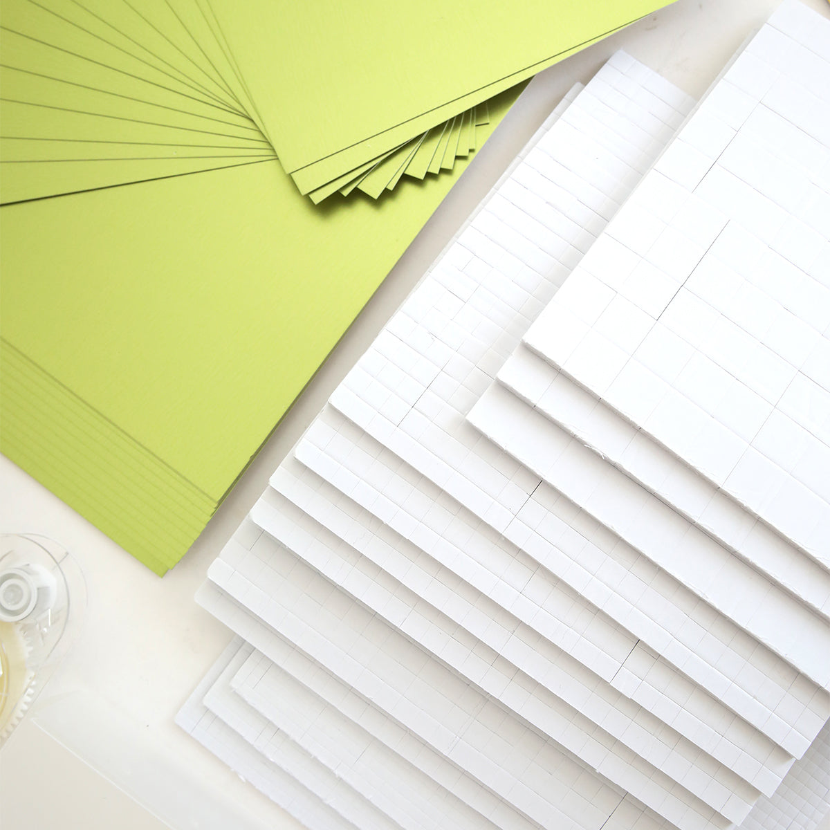 Stacks of green and white Matte Foil Cardstock from the Create Supply Box are arranged on a white surface with a roll of clear tape, ideal for making event supplies or layering under 3D Sentiment Stickers.