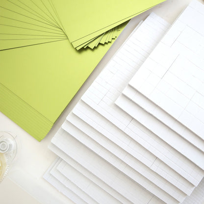 Stacks of green and white Matte Foil Cardstock from the Create Supply Box are arranged on a white surface with a roll of clear tape, ideal for making event supplies or layering under 3D Sentiment Stickers.