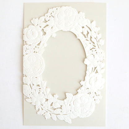 The Floral Wreath Laser Cut Invitation 10 pack features embossed white oval frames with delicate flower and leaf patterns on a pale background. The blank center is perfect for customizing your wedding invitation wraps.
