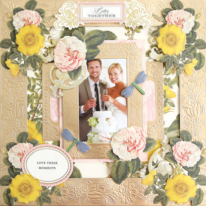 A floral-themed wedding scrapbook page highlights a beaming couple holding drinks, surrounded by decorative flowers and dragonflies. The page is enhanced with hints of Champagne Foil Matte Cardstock, adding a subtle shimmer, and the caption reads "Love These Moments.