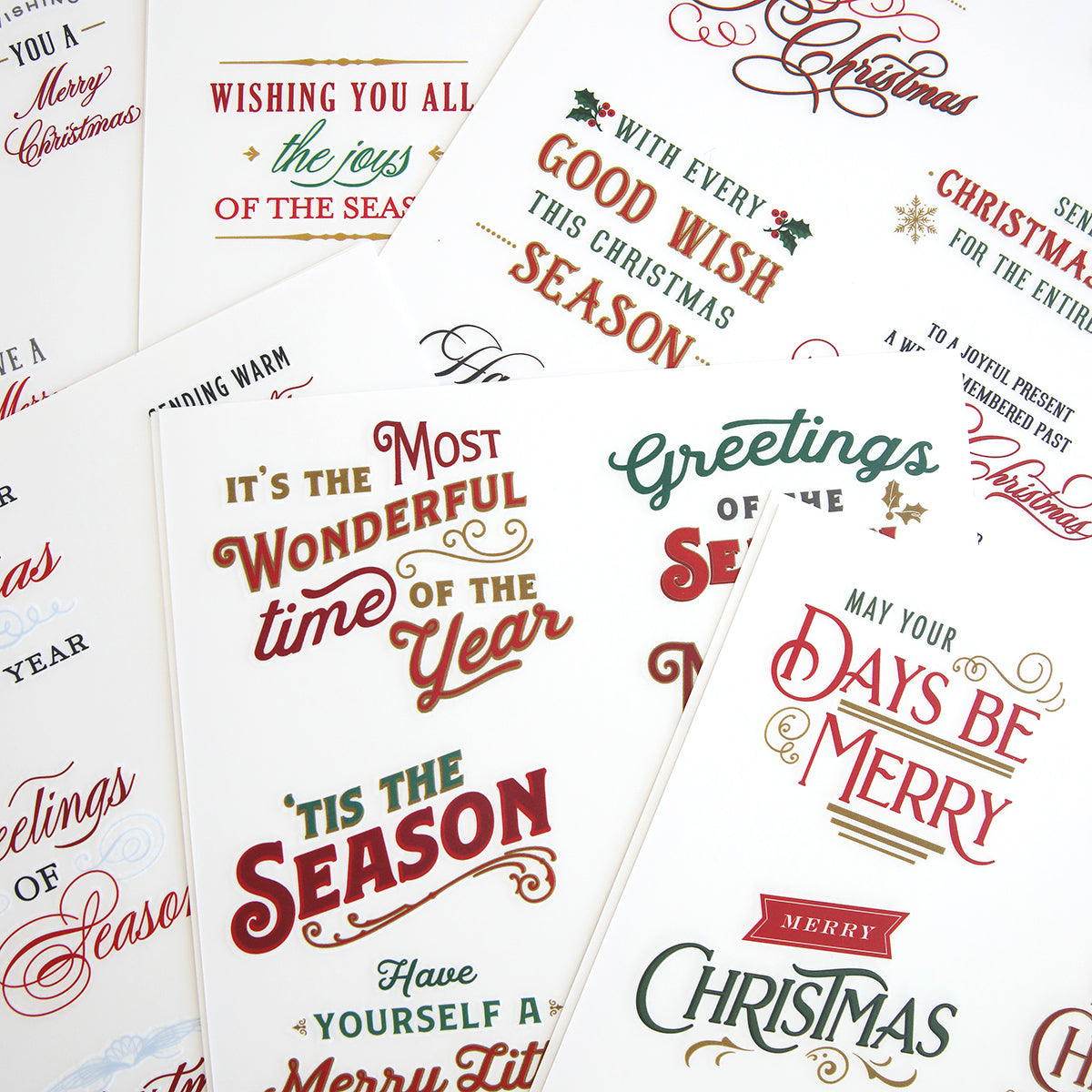 A collection of Christmas greeting cards with various festive messages, decorative fonts, and holiday-themed designs scattered across a white background, enhanced by Christmas Greetings Rub On Transfers.