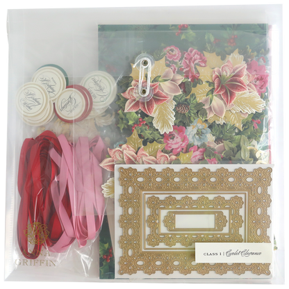 The Eyelet Elegance Dies and Materials kit includes floral paper, decorative frames, satin ribbons, seals, a metal clasp, and elegant embellishments, all neatly arranged in a clear plastic bag.
