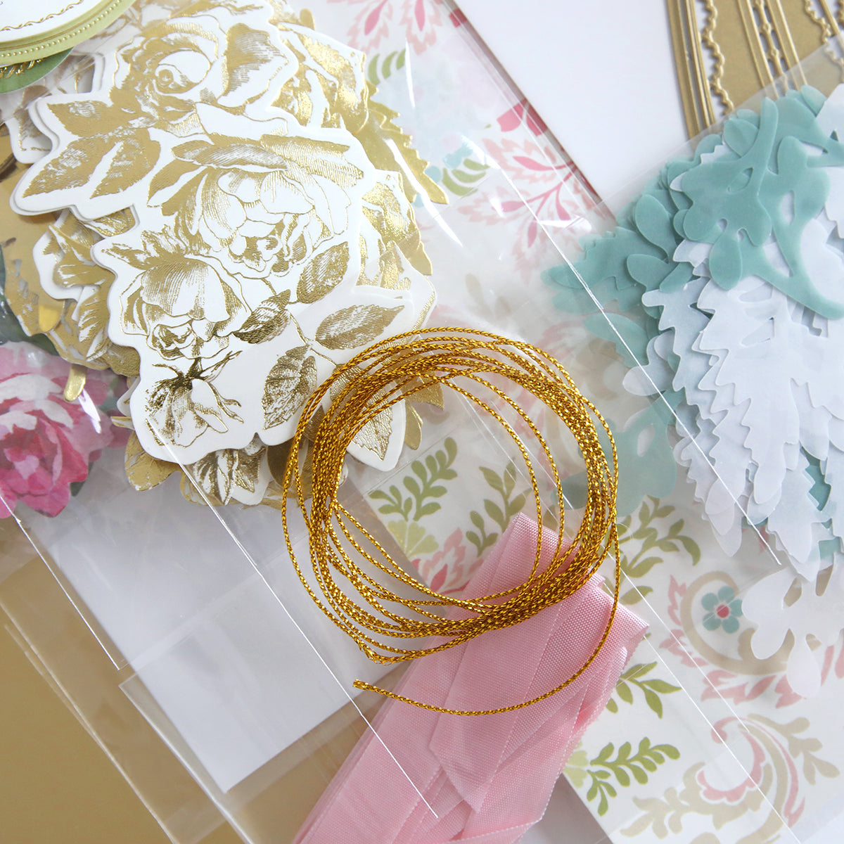 The Corset Album Dies and Materials includes decorative paper cutouts, intricate dies, a coil of gold string, and a pink ribbon exquisitely arranged on a floral-patterned background.