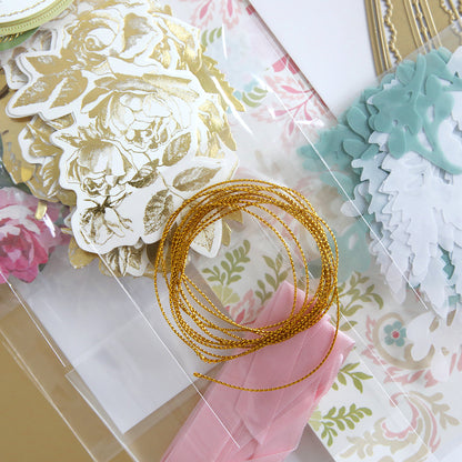 The Corset Album Dies and Materials includes decorative paper cutouts, intricate dies, a coil of gold string, and a pink ribbon exquisitely arranged on a floral-patterned background.