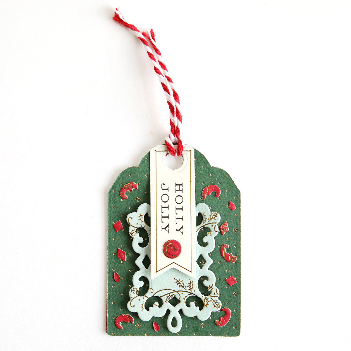 Craft festive gift or Advent Calendar tags using the Creative Tag Dies. This handmade green tag features red accents, a decorative cutout design, red and white string, and a "HOLLY JOLLY" banner in black.