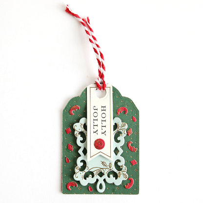 Craft festive gift or Advent Calendar tags using the Creative Tag Dies. This handmade green tag features red accents, a decorative cutout design, red and white string, and a "HOLLY JOLLY" banner in black.