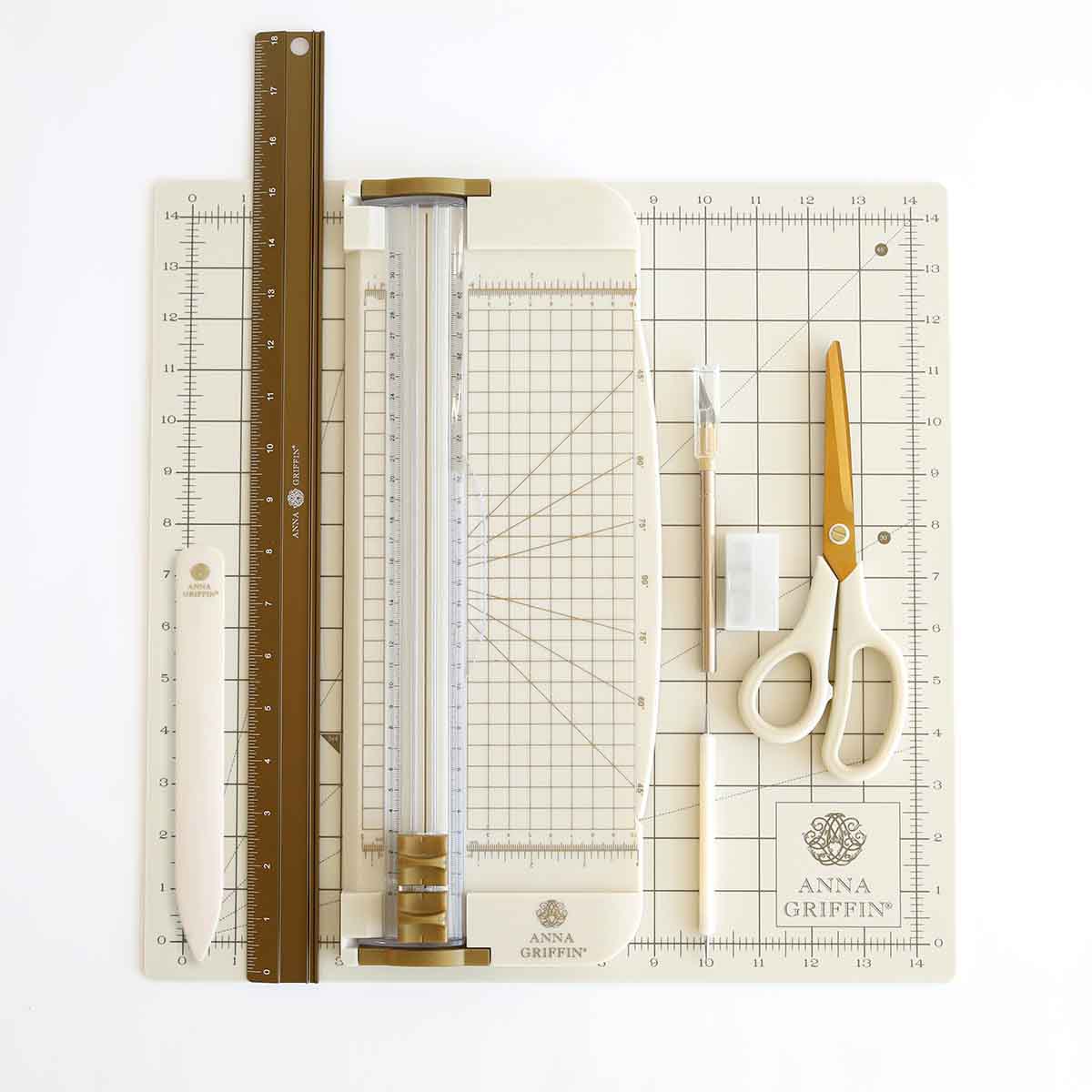 The Essential Tool Set—featuring a ruler, bone folder, paper trimmer, scissors, utility knife, and cutting mat—is neatly arranged on a gridded Anna Griffin-branded surface for card making and other crafts.
