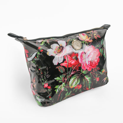The Noire Large Cosmetic Bag is a glossy black zippered pouch adorned with a floral pattern of pink and white flowers and green leaves, accented by a round metal emblem on the front. Ideal as a spacious cosmetic bag or travel organizer, it keeps your essentials stylishly organized wherever you go.