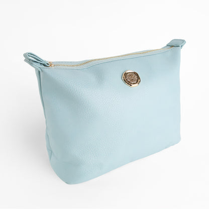 A Beach Glass Blue Large Cosmetic Bag made from faux leather, featuring a gold zipper and a round gold emblem on the front, is photographed against a plain white background. Combining style with practicality, this spacious cosmetic pouch is perfect for travel.