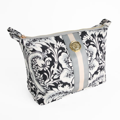 The Damask Noire Large Cosmetic Bag features a black and white floral design with beige and gray stripes, accented by a round gold emblem on the front. Displayed on a white background, this stylish large makeup bag is perfect for organizing your essentials at home or on the go.