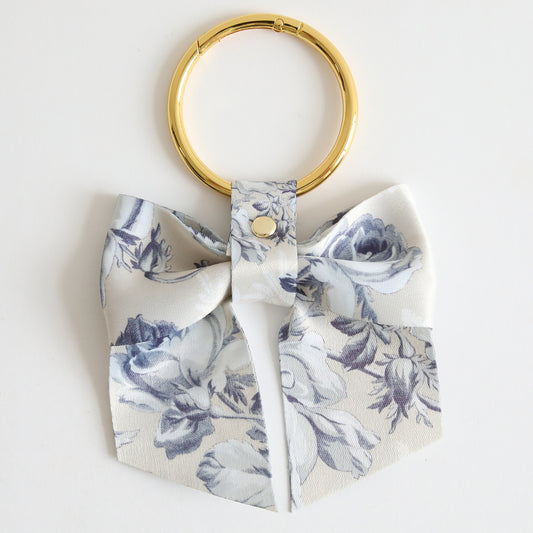 The Faded Rose Bag Charm features a gold keyring with a white twill bow adorned in a blue floral pattern, secured by a metal rivet and finished with a gold clasp—an elegant accessory to enhance any bag.