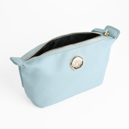 The Beach Glass Blue Small Cosmetic Bag is a light blue faux leather zippered pouch featuring a gold emblem on the front, displayed open to reveal its black interior lining against a white background—perfect as a stylish travel cosmetic pouch or small makeup bag.