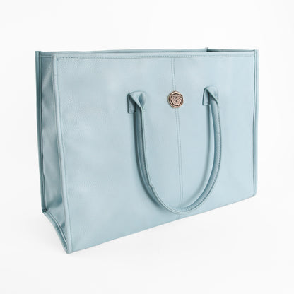 The Beach Glass Blue Tote Bag is a light blue, rectangular tote crafted from premium faux leather. It features two handles and a circular gold emblem on the front. This large shoulder bag is displayed against a plain white background.