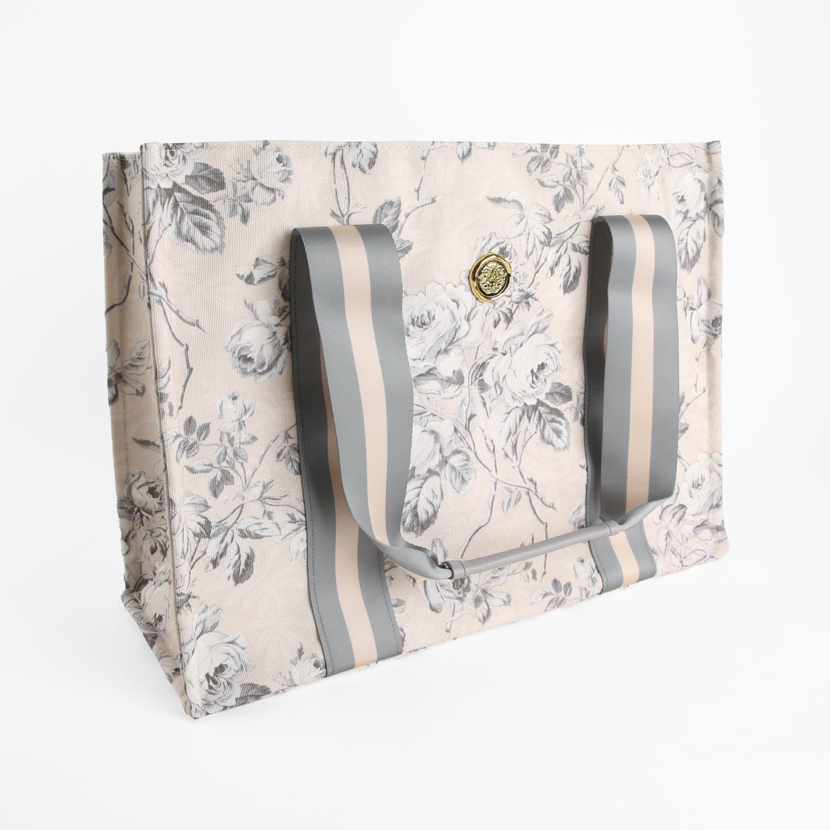 The Faded Rose Tote Bag is a large rectangular canvas tote featuring a chic gray and beige floral pattern, wide striped handles, and a gold circular emblem on the front—making it the perfect stylish market bag for women.