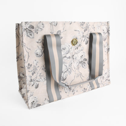 The Faded Rose Tote Bag is a large rectangular canvas tote featuring a chic gray and beige floral pattern, wide striped handles, and a gold circular emblem on the front—making it the perfect stylish market bag for women.