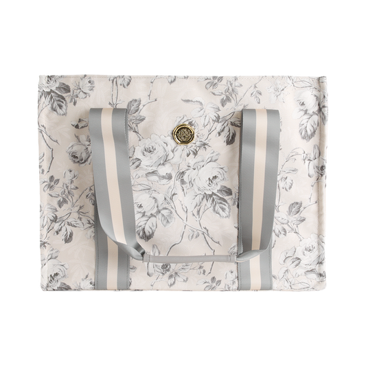 The Faded Rose Tote Bag features a rectangular canvas design with a gray and white floral print, double gray striped handles, and a round gold emblem on the front—making it the perfect stylish floral market bag.