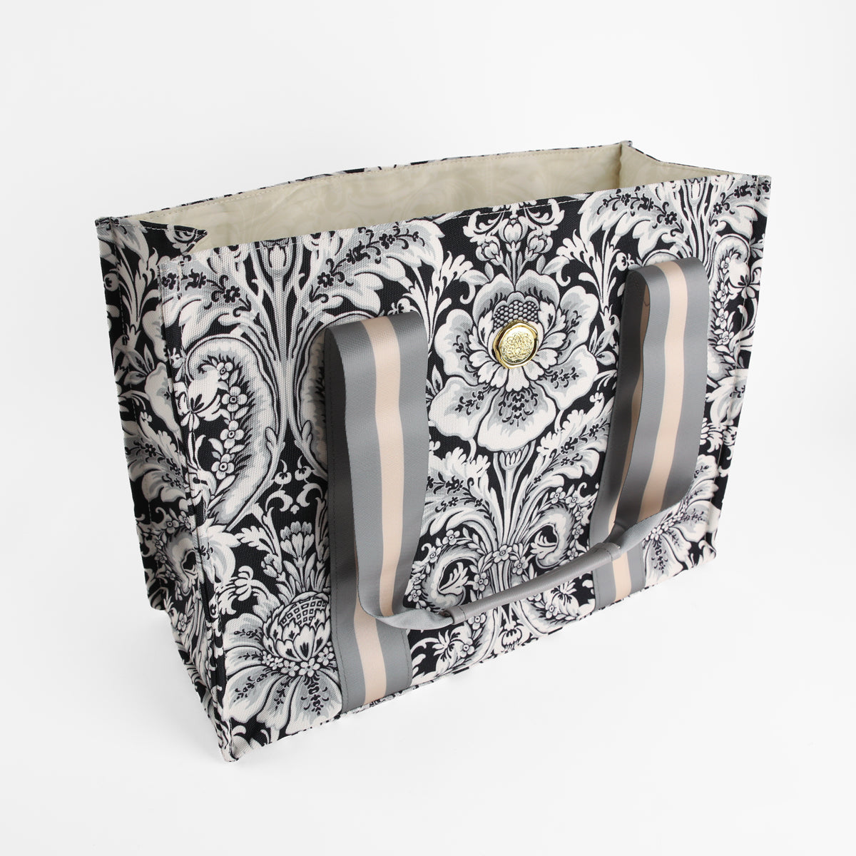 The Damask Noire Tote Bag is a large, rectangular canvas tote featuring a striking black and white damask floral pattern, a beige interior, striped handles, and a circular gold emblem on the front—an elegant gift for women or the perfect floral market bag.