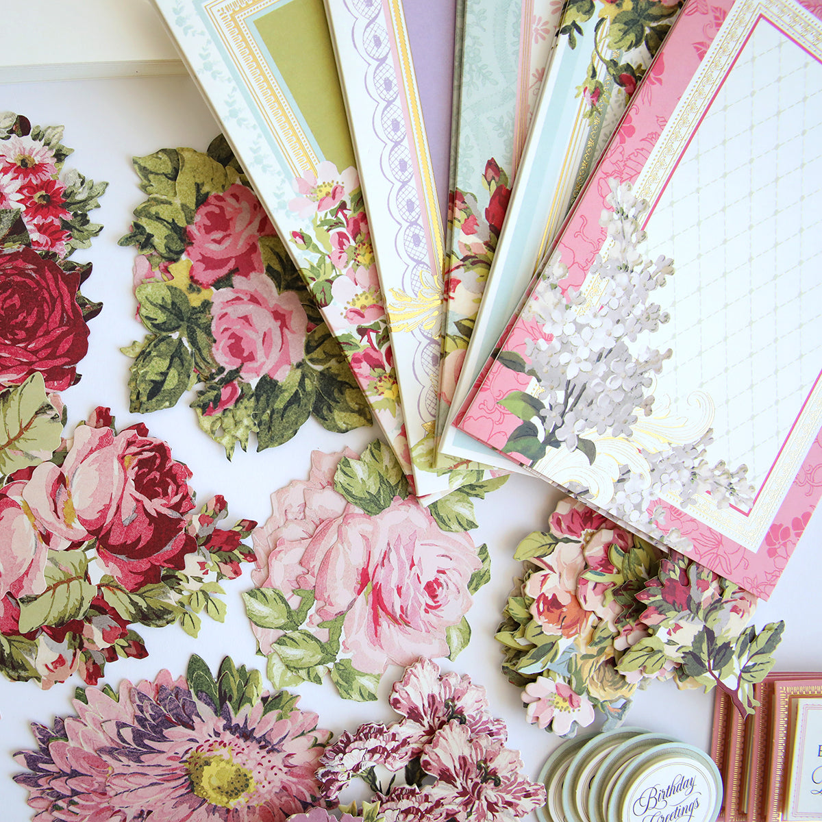 Various floral patterned scrapbooking papers and cut-out flowers from the Folded Flower Birthday Card Kit scattered on a white surface.